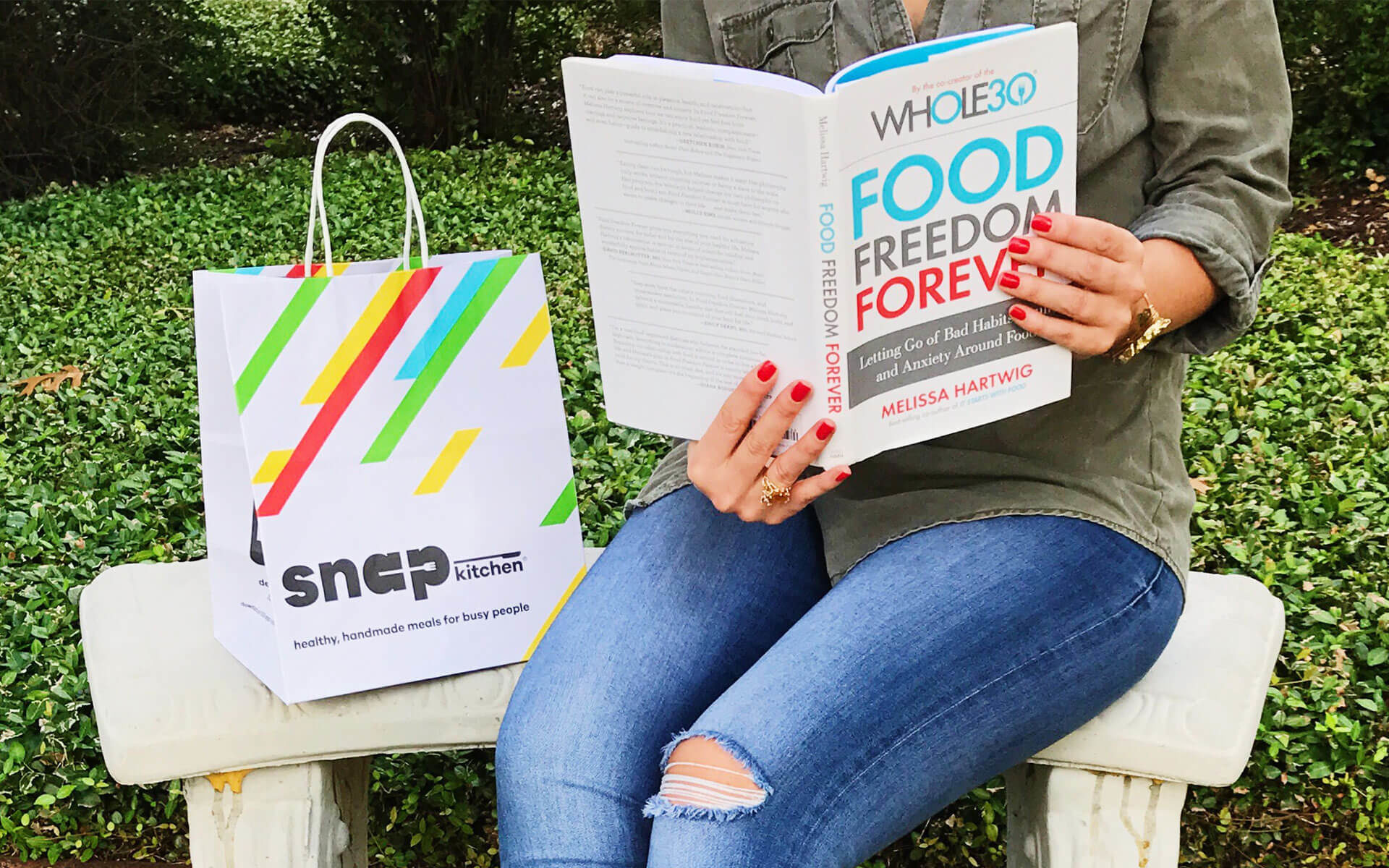woman sitting on a bench next to a Snap Kitchen bag reading Food Freedom Forever book