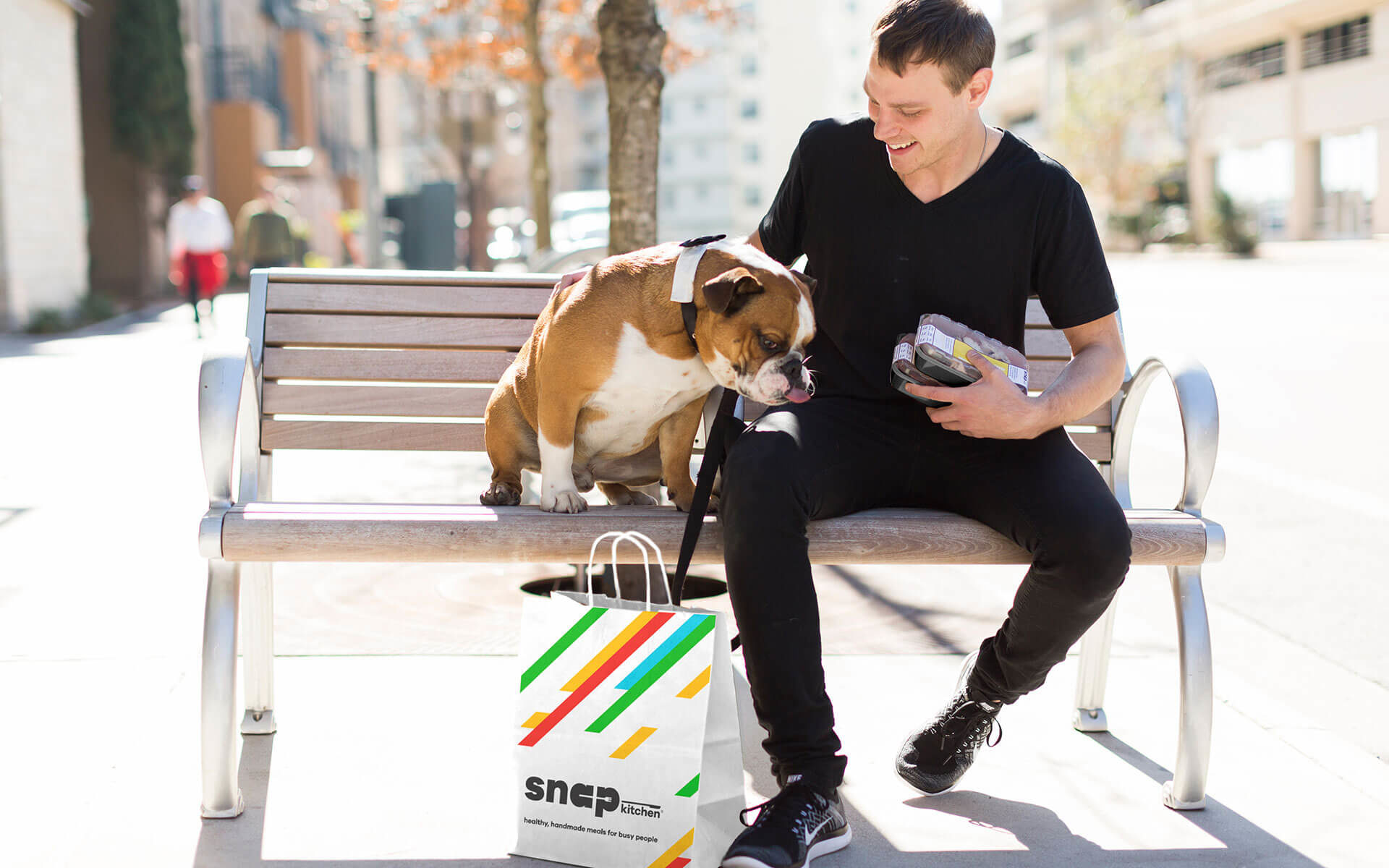 a man and a bulldog sitting on a bench with a bag from Snap Kitchen