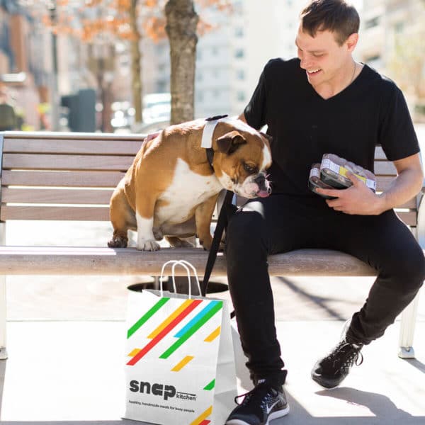 a man and a bulldog sitting on a bench with a bag from Snap Kitchen