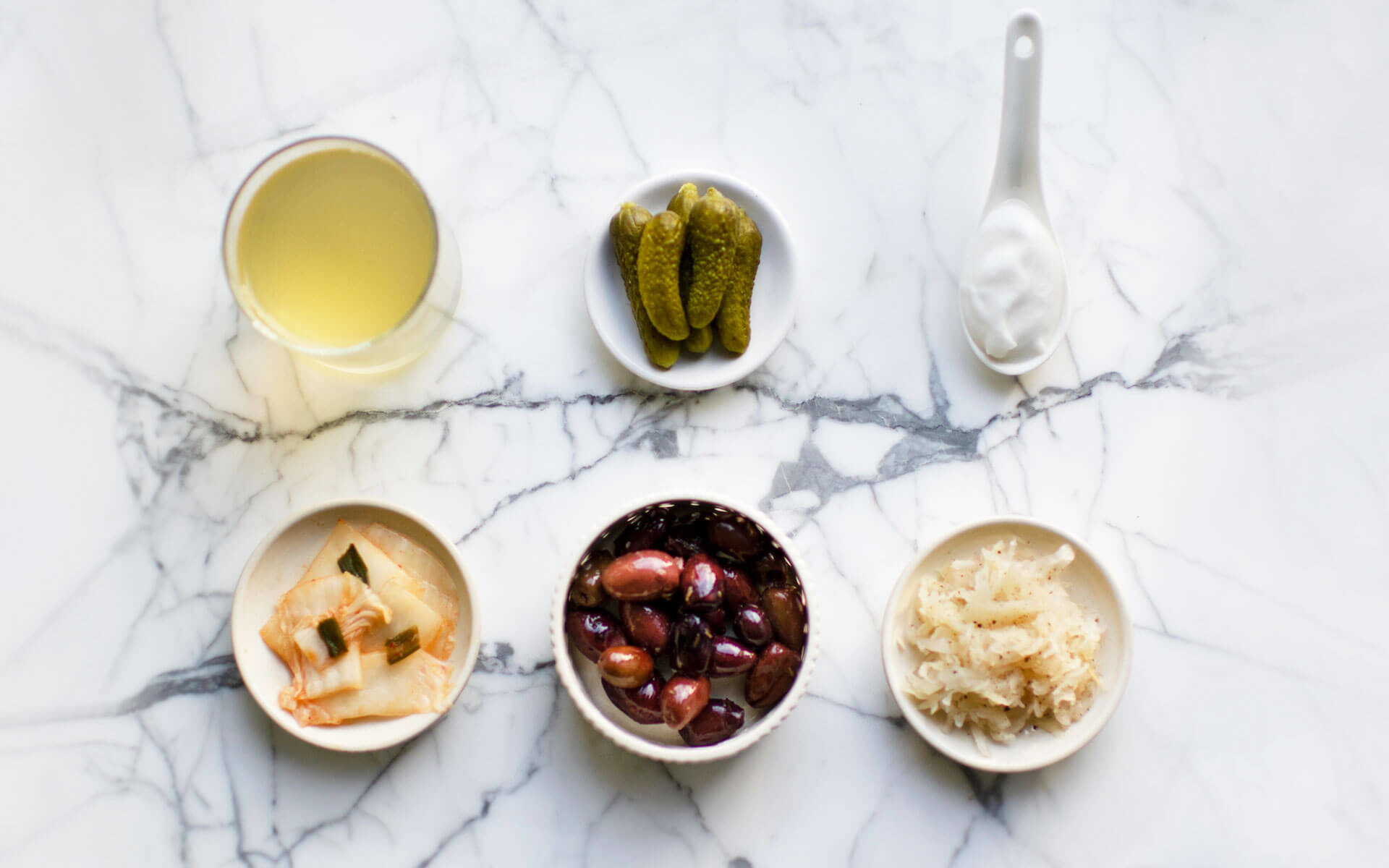 vinegar, pickles, yogurt, kimchi, olives and sauerkraut in bowls on a marble table