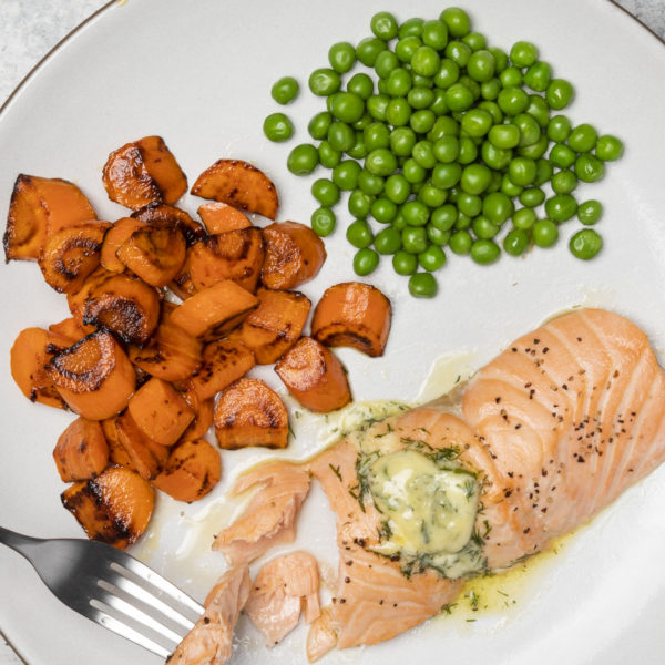 Salmon, carrots and peas on a plate. There is a hand and fork visible picking up a piece of the salmon
