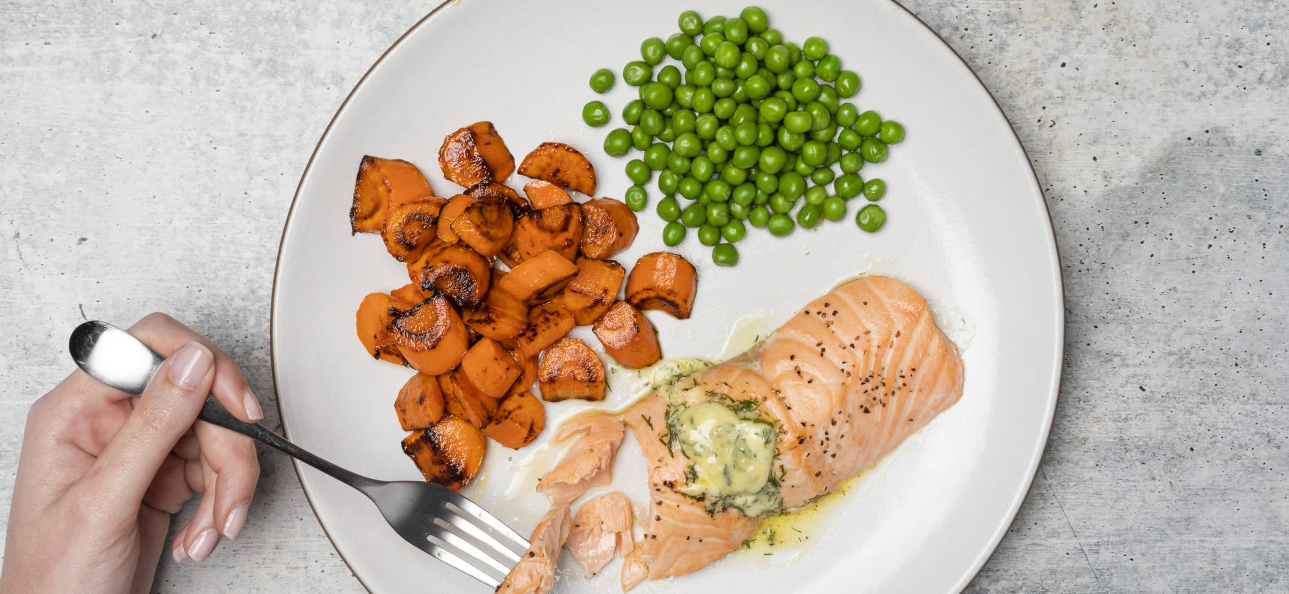 Salmon, carrots and peas on a plate. There is a hand and fork visible picking up a piece of the salmon