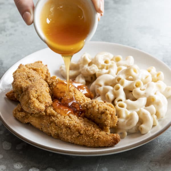 Hot honey being poured over breaded chicken on a plate with mac and cheese on the side