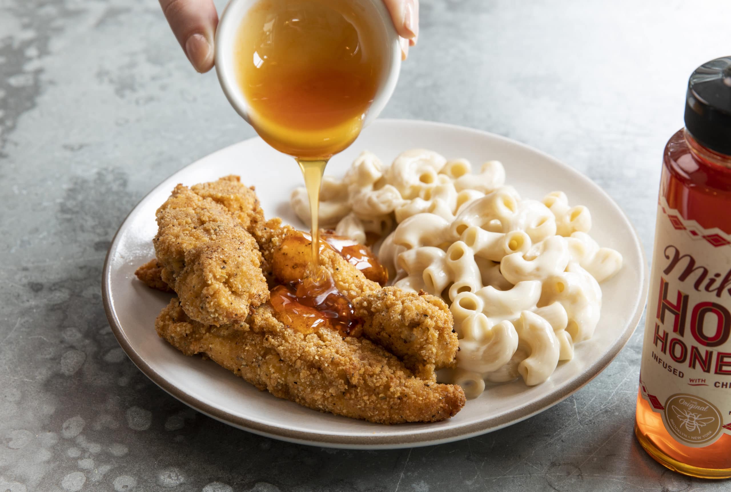 Hot honey being poured over breaded chicken on a plate with mac and cheese on the side