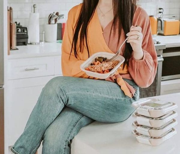 Woman with long dark hair and an orange top, sitting on a white counter eating out of a to-go container