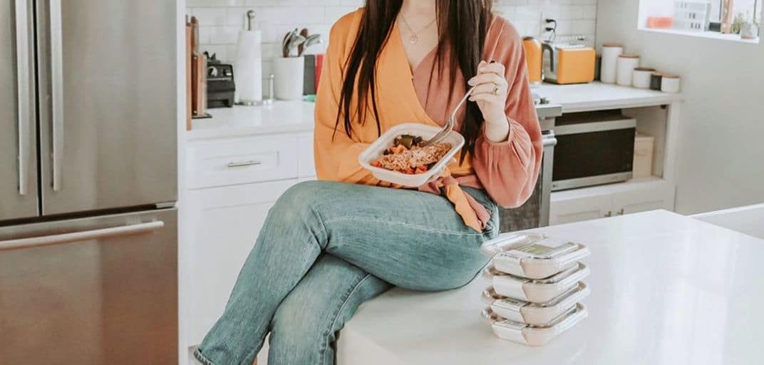 Woman with long dark hair and an orange top, sitting on a white counter eating out of a to-go container