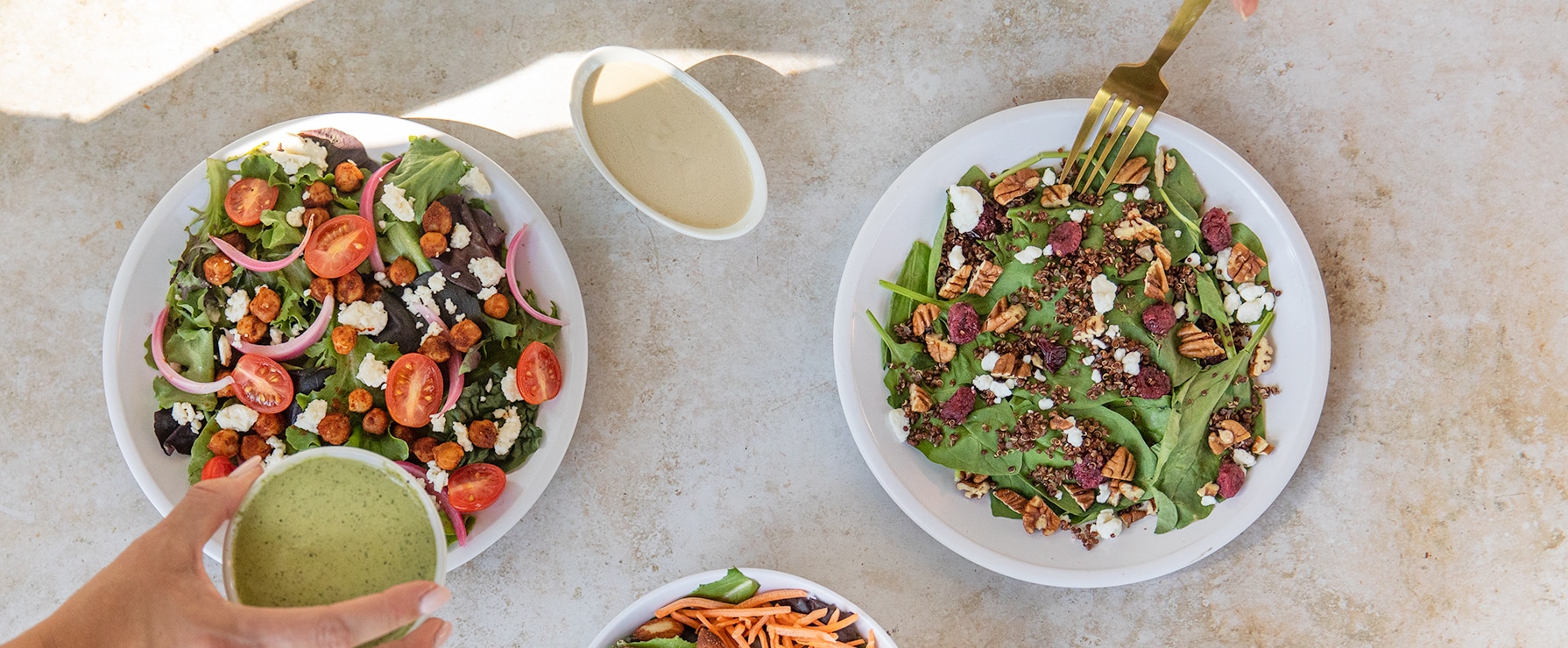 Two plates of salad, one with tomatoes and one without, on a marble countertop with different dressings