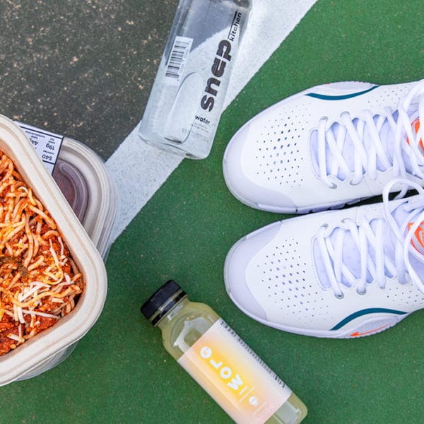 A pair of white sneakers with a tennis ball sitting inside of them, with bottles of water and juice, and a to-go container of spaghetti sitting on pavement