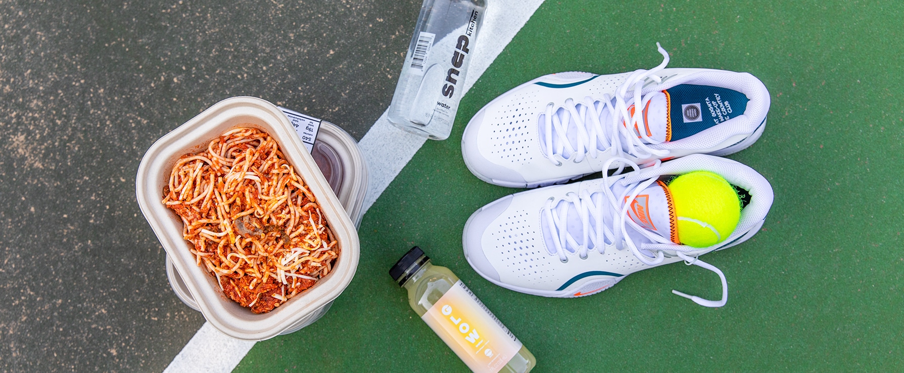 A pair of white sneakers with a tennis ball sitting inside of them, with bottles of water and juice, and a to-go container of spaghetti sitting on pavement