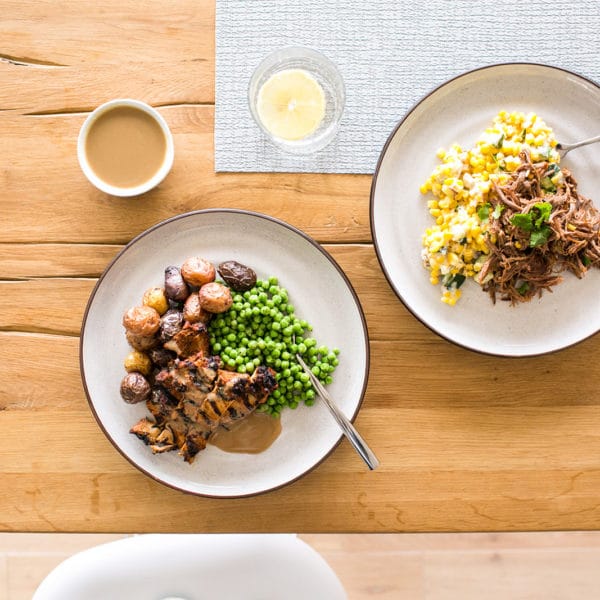 Two plates on a wood table - one with chicken, peas and potatoes, and another with pulled pork and corn.