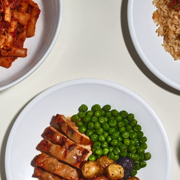 Three plates of food on a white background, one with pasta, one with chicken, potatoes and peas, and the third with rice, chicken and tomatoes.