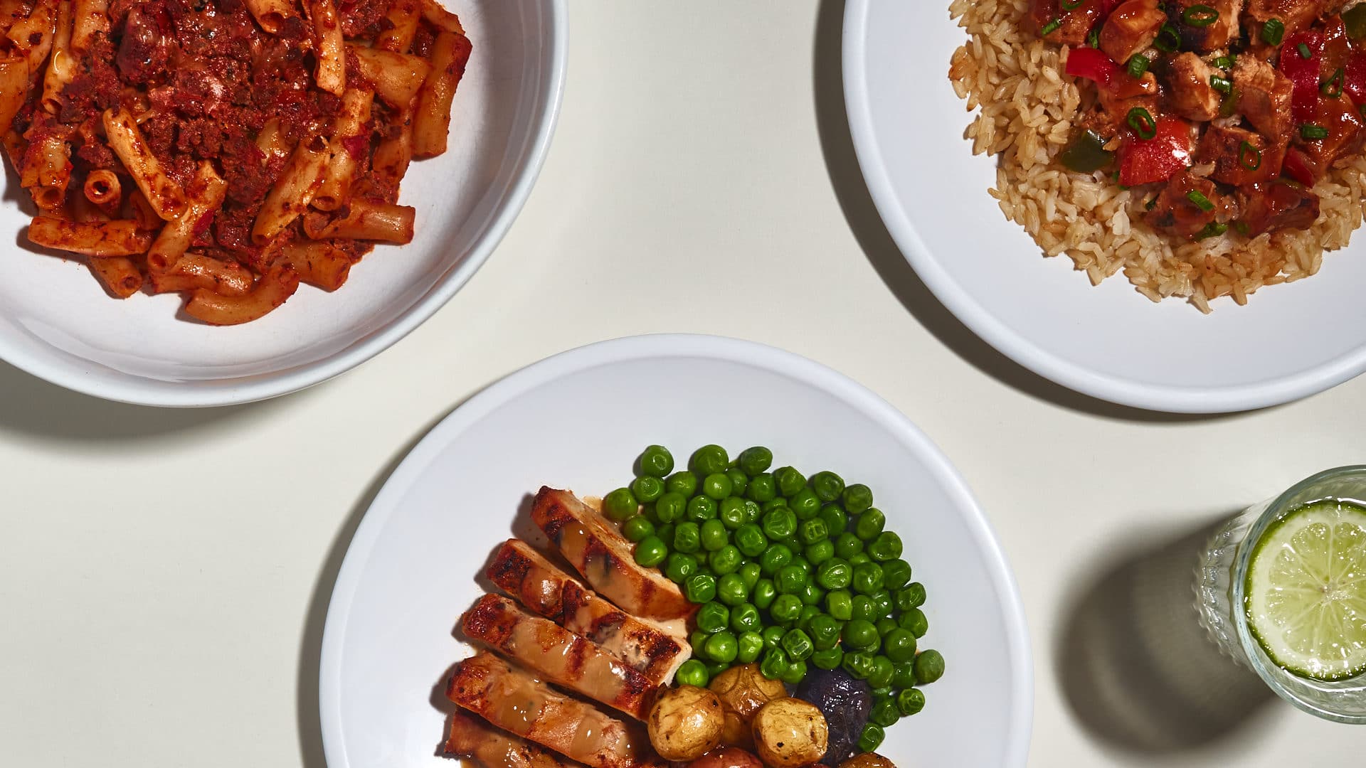 Three plates of food on a white background, one with pasta, one with chicken, potatoes and peas, and the third with rice, chicken and tomatoes.