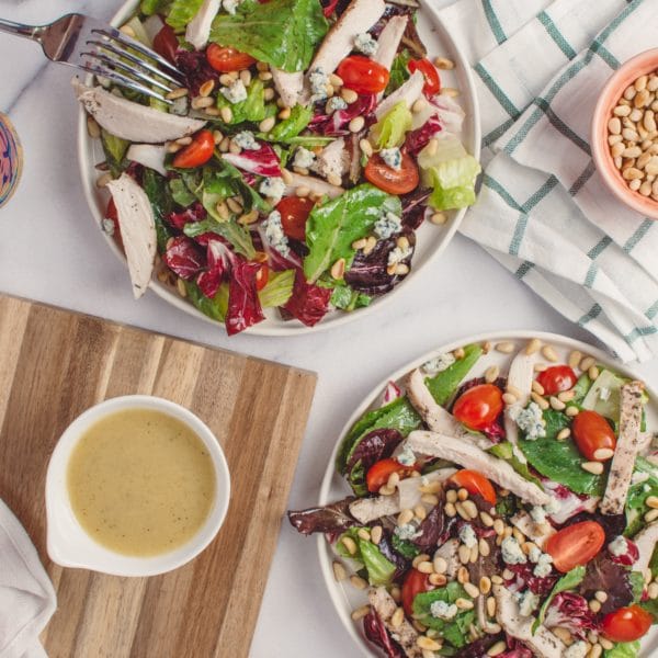 Two salads with chicken on a white background alongside a bowl of dressing, a fork and a can of seltzer