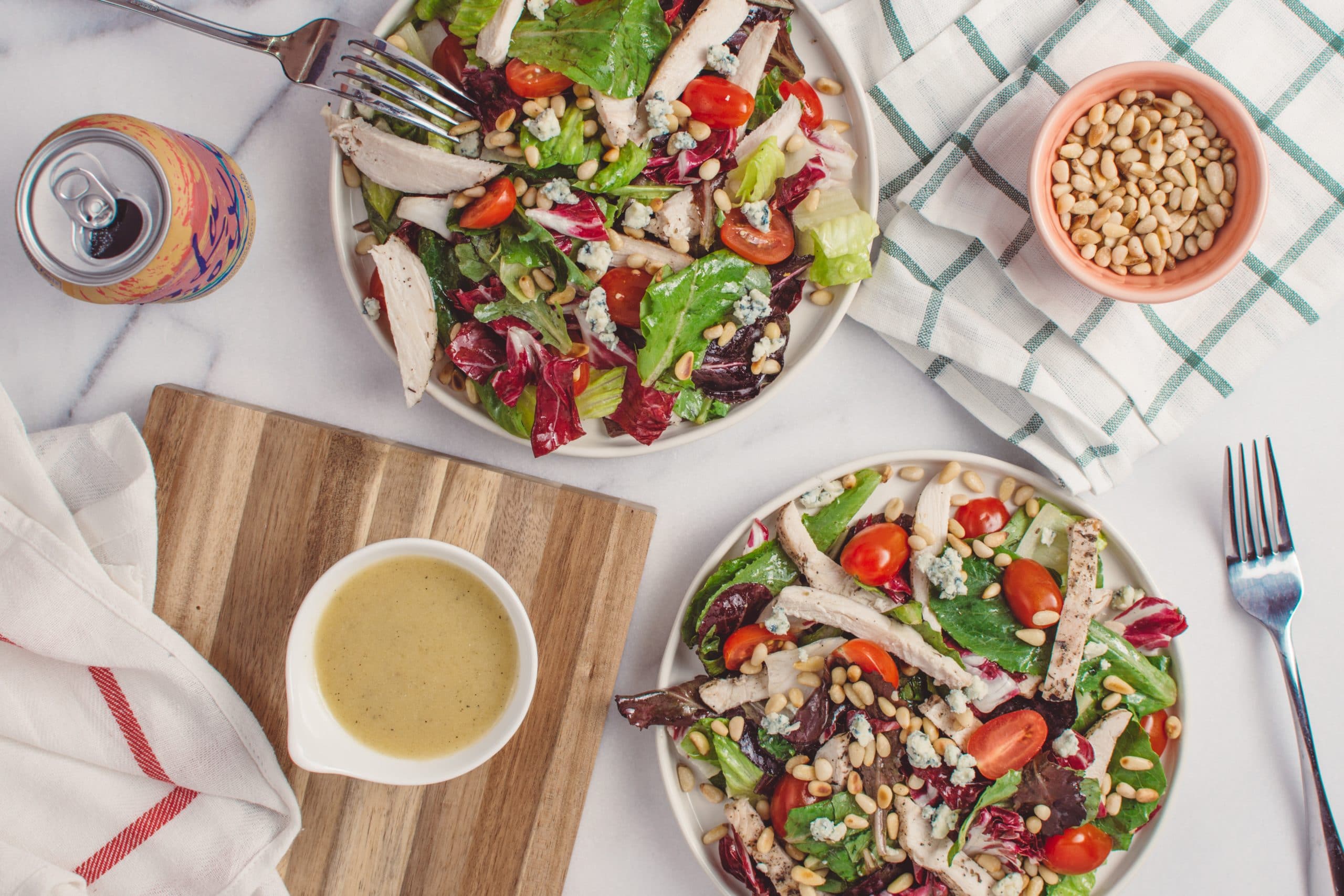 Two salads with chicken on a white background alongside a bowl of dressing, a fork and a can of seltzer