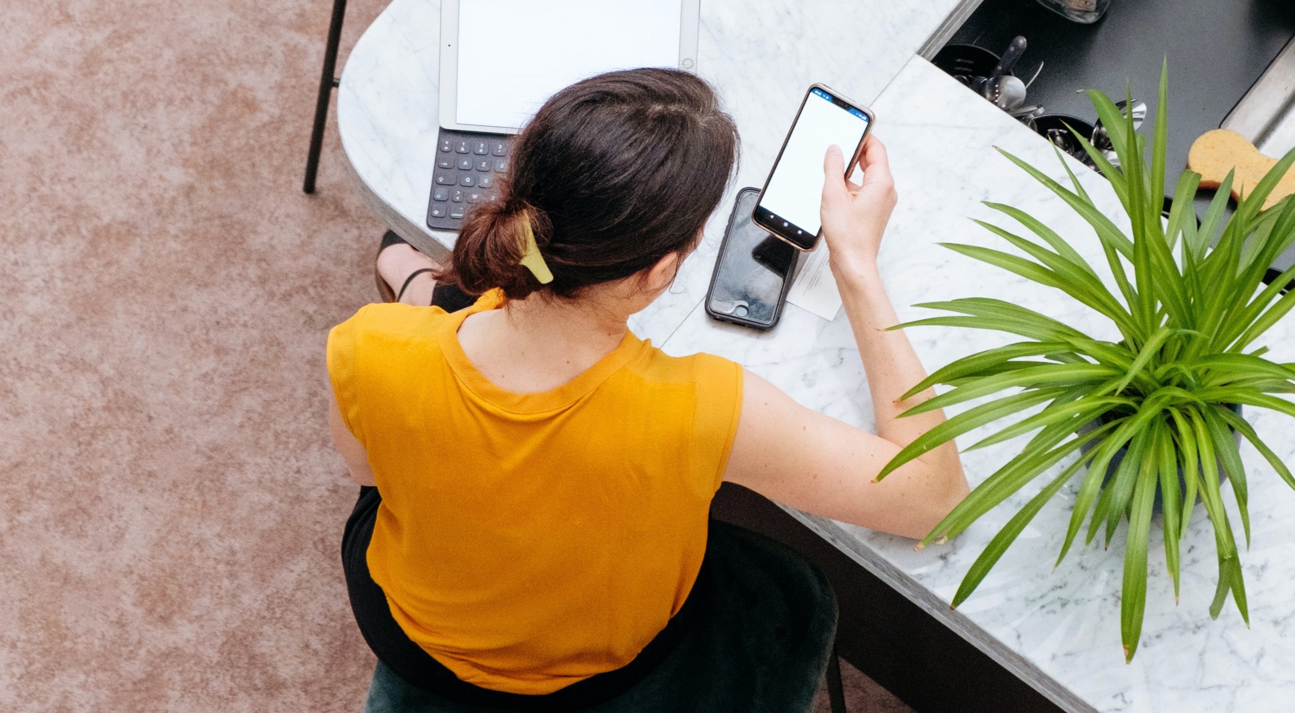 Woman in a yellow shirt sitting at a desk with a laptop, looking at her phone