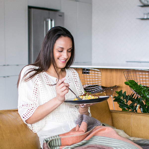 Woman with dark hair sitting on a tan couch with a plate of food and smiling