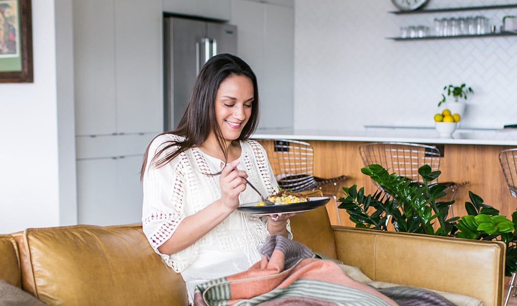 Woman with dark hair sitting on a tan couch with a plate of food and smiling