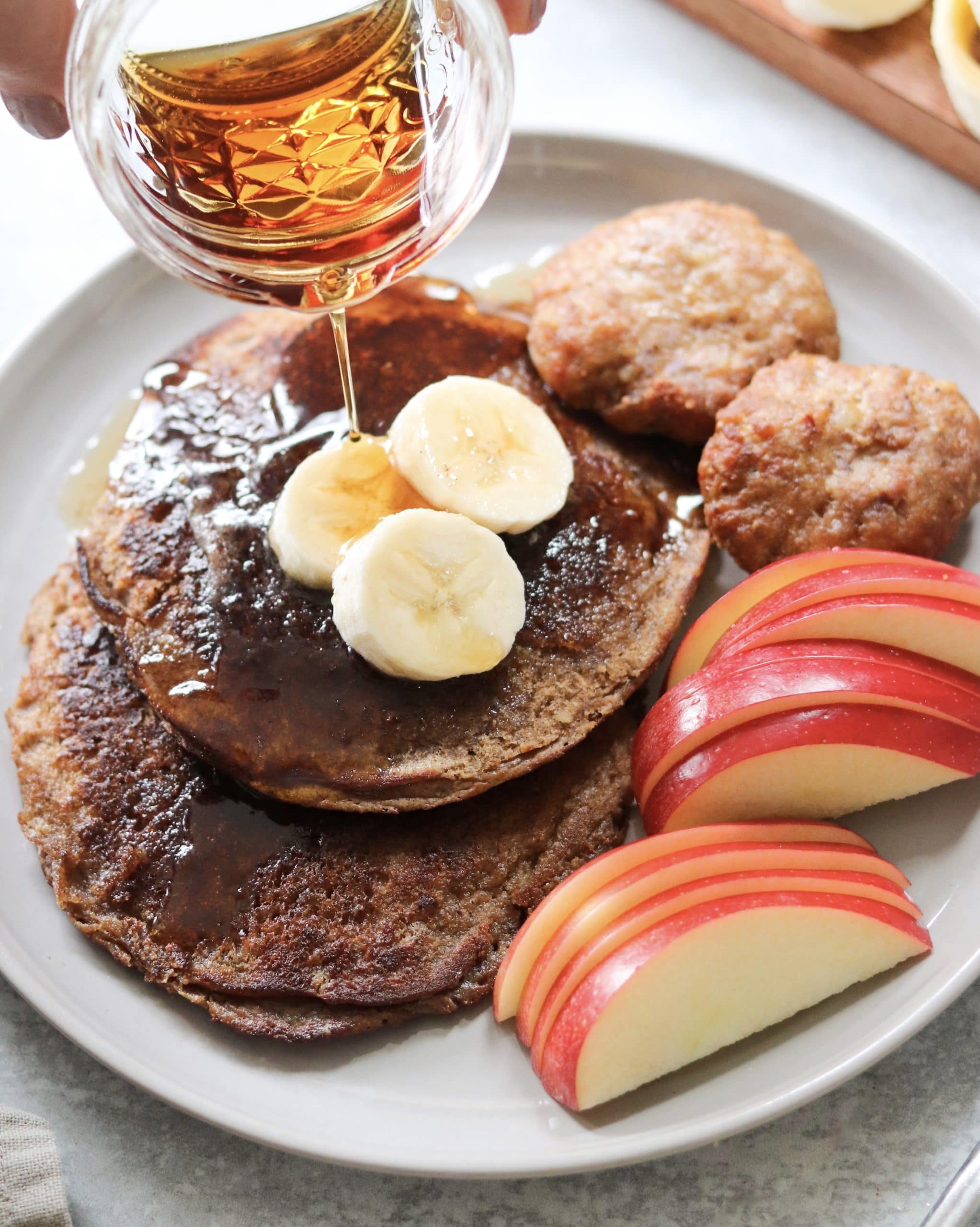 Syrup being poured over a plate of pancakes with apples and bananas