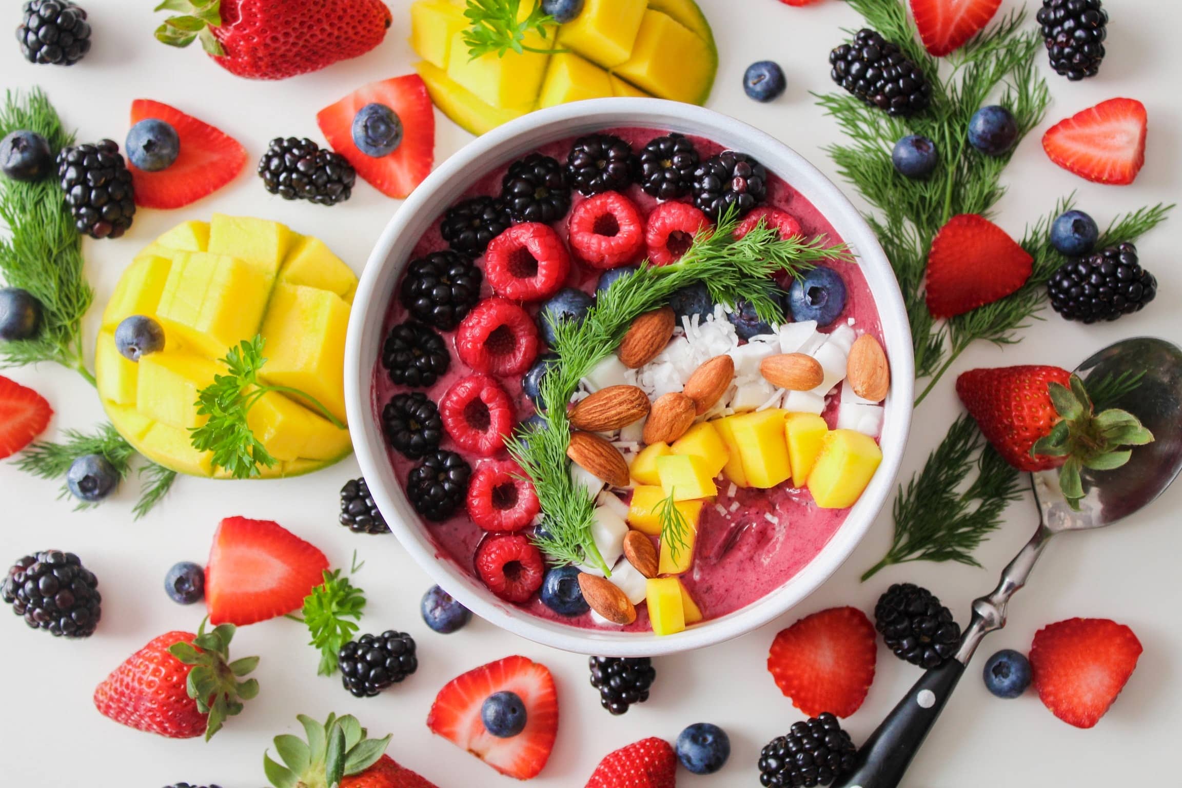 Smoothie bowl with a variety of berries, pineapple, almonds, and coconut. The smoothie bowl is surrounded by fruit.
